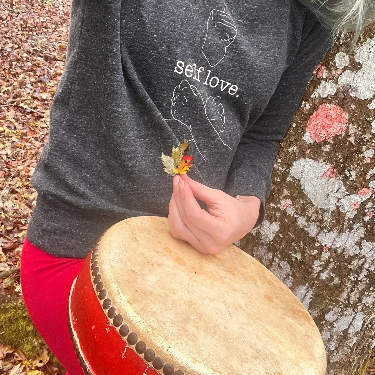 A woman in a “self love” sweatshirt holds a small autumn leaf above a hand drum.