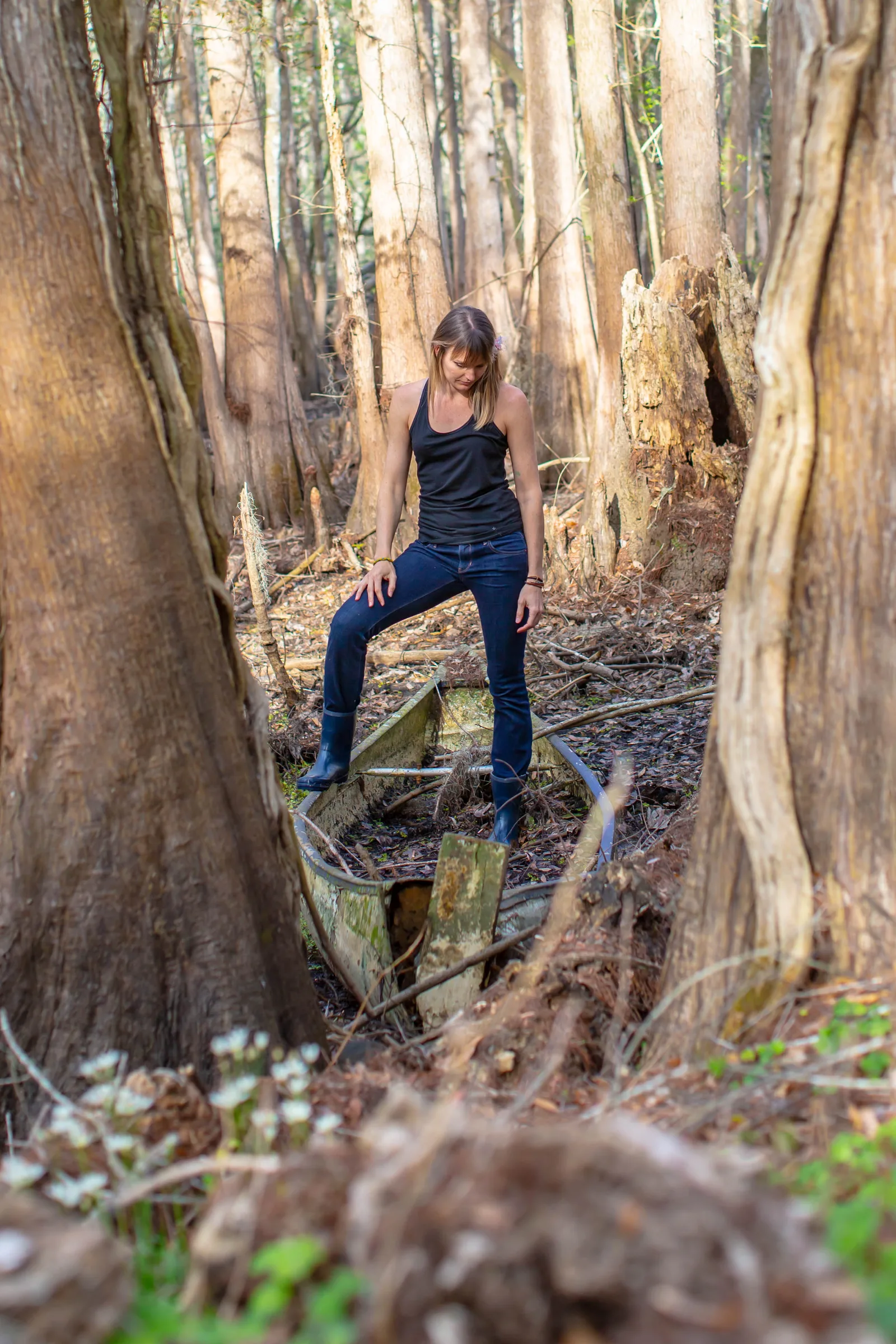 A woman stands on an old wooden boat among cypress trunks in a swamp forest.