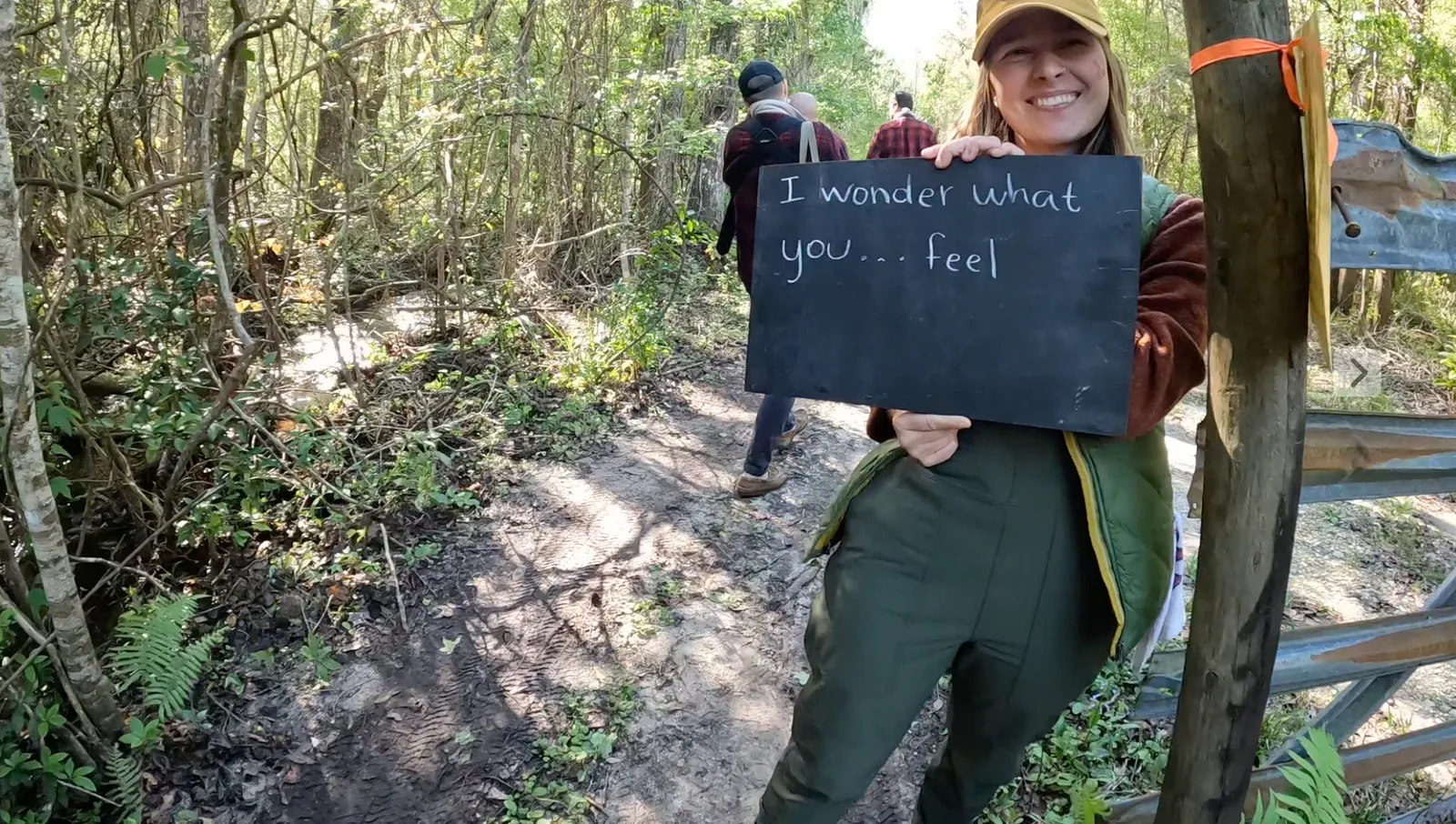 A smiling guide on a forest trail holds up a chalkboard that reads “I wonder what you… feel.”