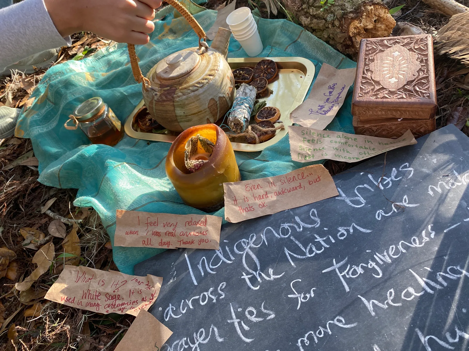 A teapot, sage and handwritten notes laid on a cloth beside a chalkboard with a message about Indigenous tribes, forgiveness and healing.