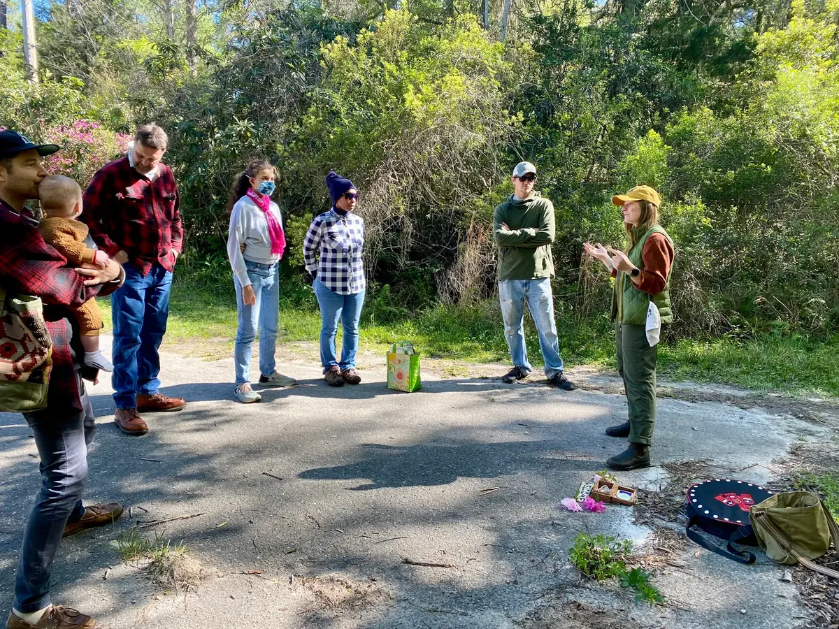 A group stands in a loose circle on a path while a guide in a green vest and cap speaks to them.