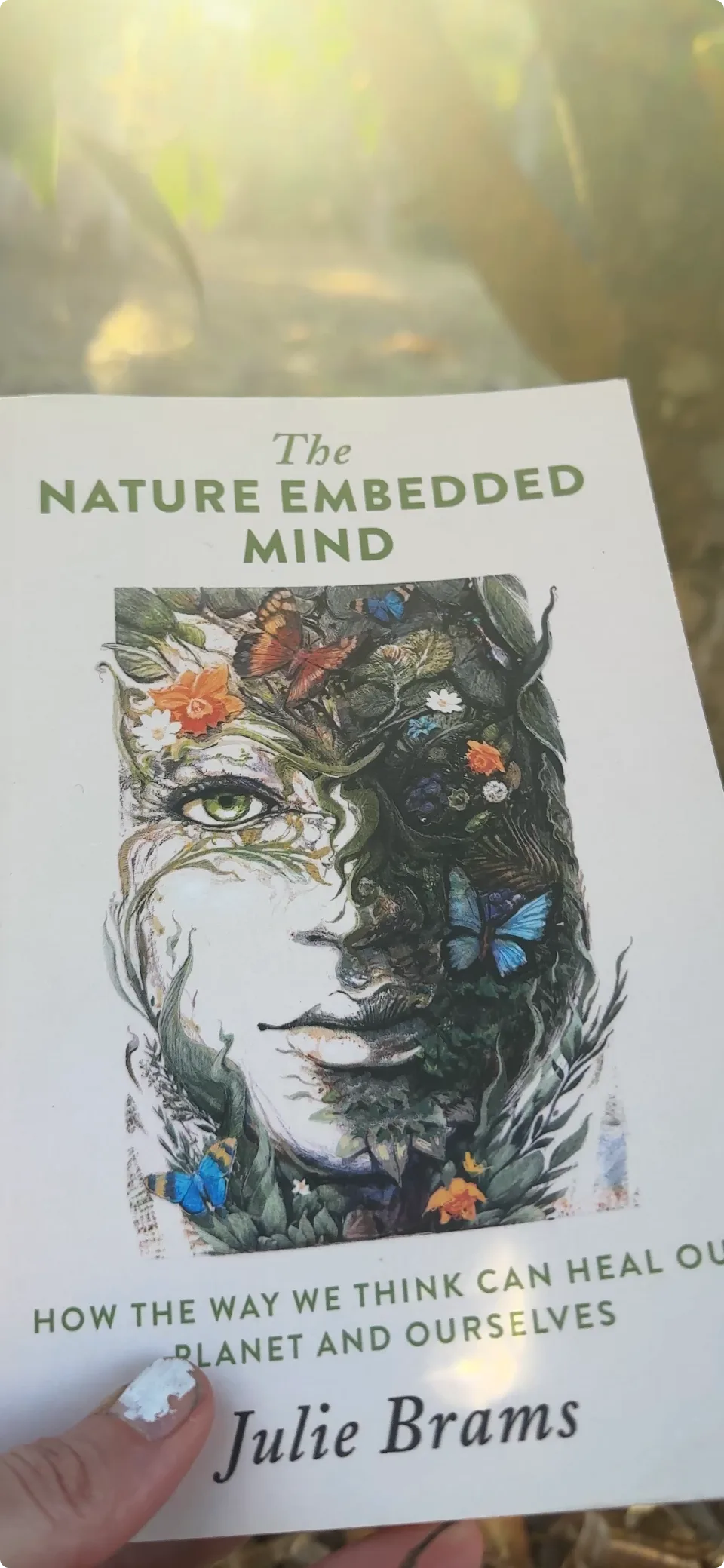 A hand holds the book The Nature Embedded Mind by Julie Brams in the sunlight.