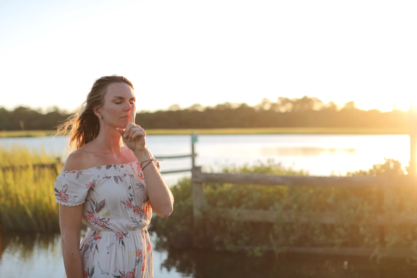 A woman in a floral dress stands by the water at sunset, eyes closed, a finger to her lips.