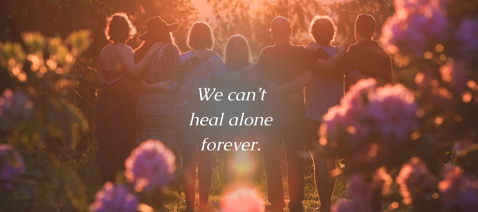 [image description: Group of people standing shoulder-to-shoulder, arms around each other, watching a warm sunset. Pink flowers frame the foreground. White text overlay reads, ‘We can’t heal alone forever.’ Atmosphere is calm and peaceful"]