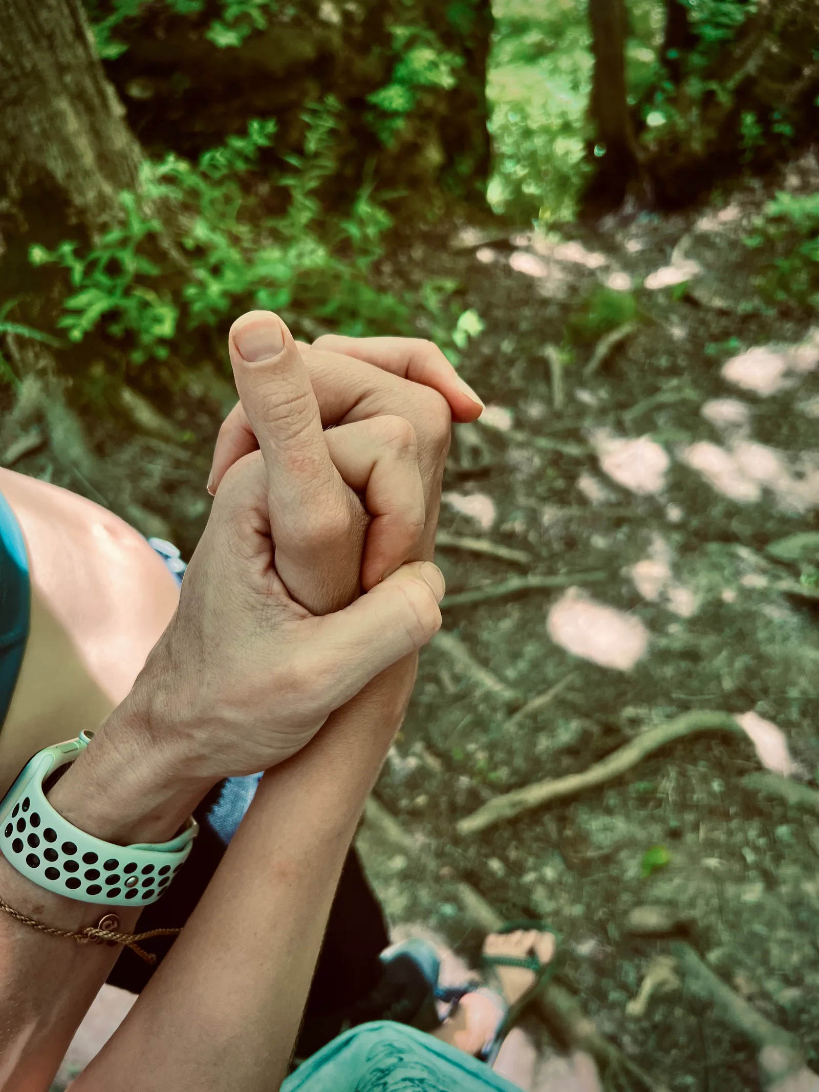 [id: Summer and Meredith’s hands intertwine, their feet and a patch of woods blurry in the background]