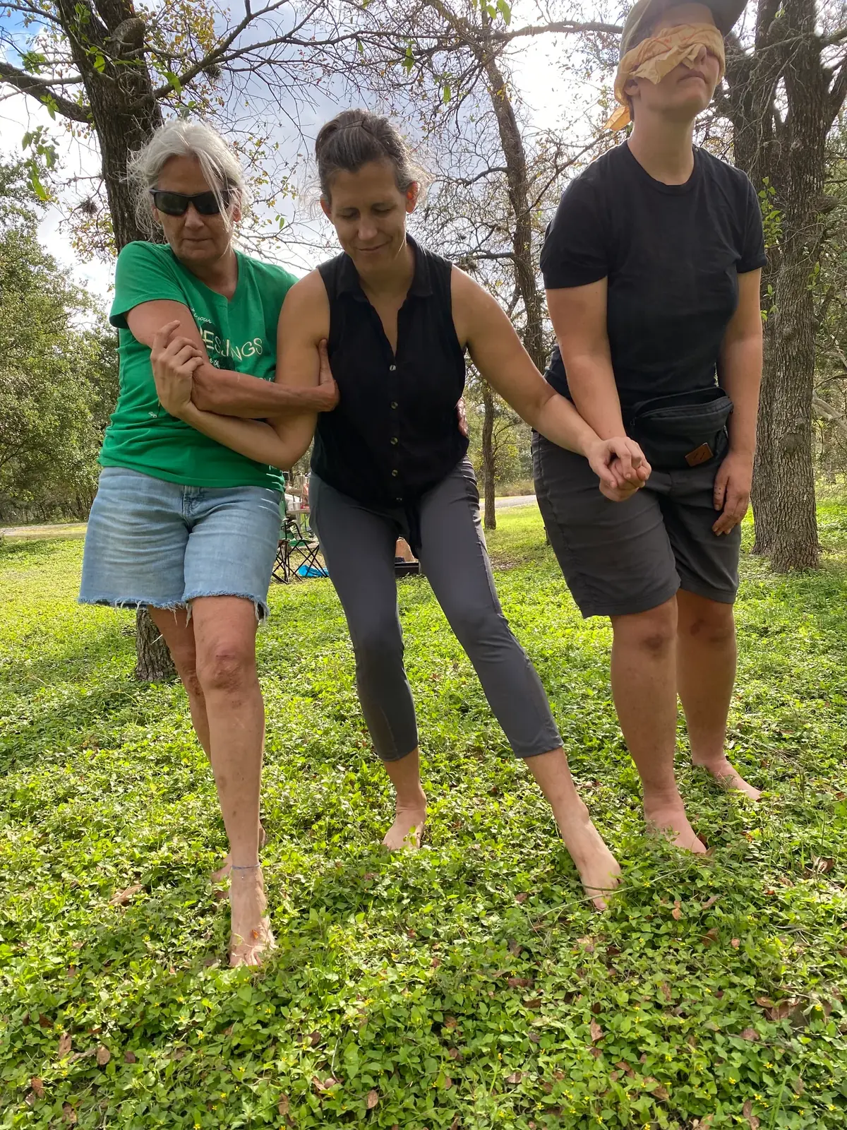 [id: Three individuals, one blindfolded and the others’ eyes closed, embrace one another and walk barefoot on soft, herb-covered ground]