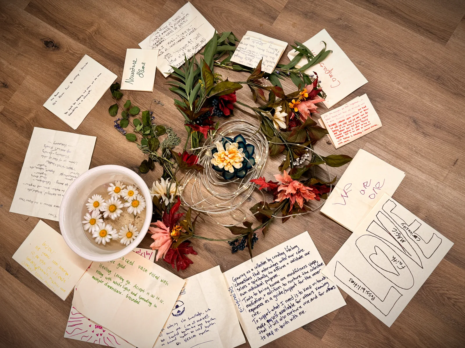 Handwritten notes and cards laid in a circle around flowers and a bowl of daisies on a wooden floor.