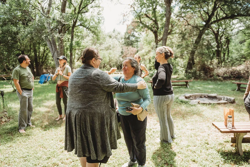 People hug and talk on a lawn under trees, near a fire ring and picnic tables.