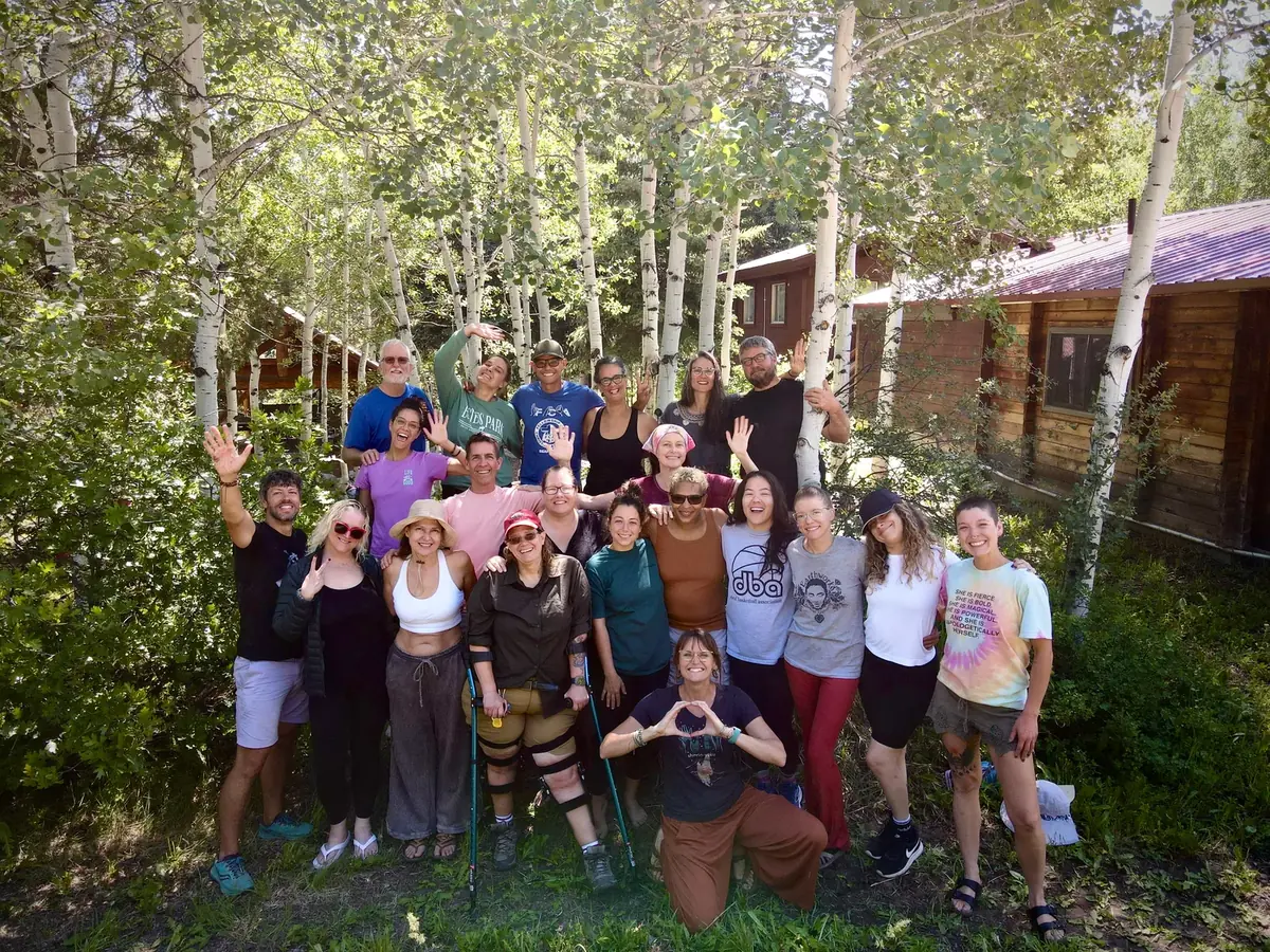 A large group of retreat participants smile and wave among aspen trees beside a log cabin.