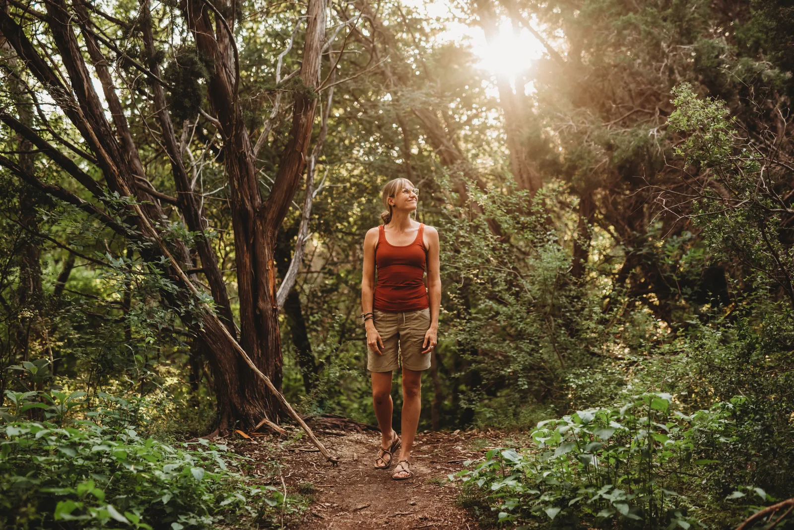 A woman wearing red tanktop and a hiker short walking in a sunlit forest, looking calmly with a peaceful smile.